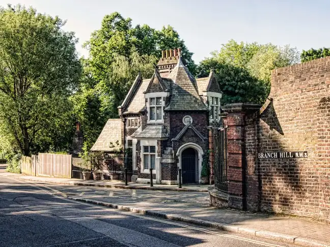 Victorian brick lodge with a steep slate roof and decorative chimneys beside a road